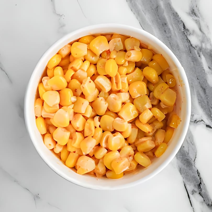 A white bowl filled with golden corn kernels, lightly seasoned, sits on a white marble surface.